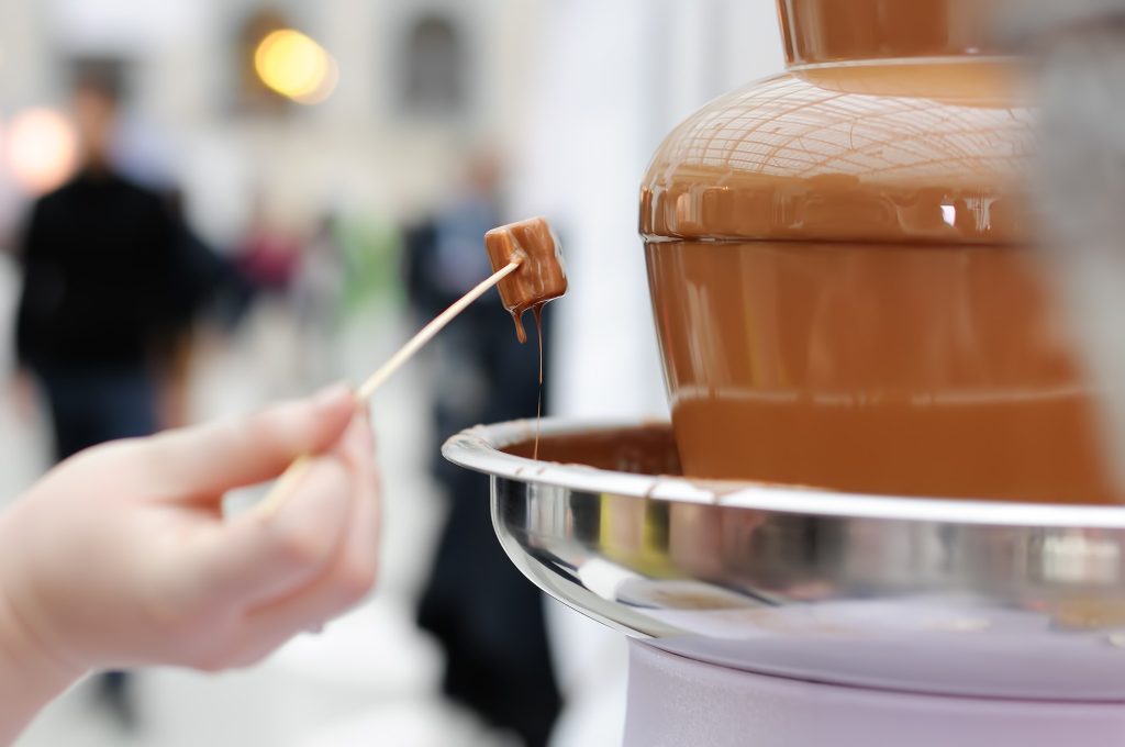 Woman holding marshmallow and frosting its in chocolate fondue fountain.